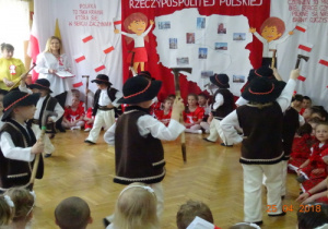Na tle dekoracji z mapą Polski i dziećmi z chorągiewkami przedszkolaki przebrane za górali tańczą z ciupagami.